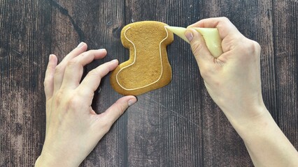 Hands skillfully decorate gingerbread cookies in the shape of a Christmas stocking with icing on a wooden table.
