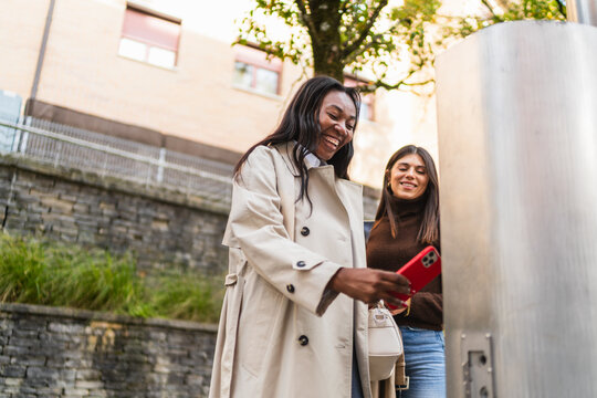 Happy women friends using a mobile phone to make a secure contactless payment at an outdoor terminal in the city - Powered by Adobe