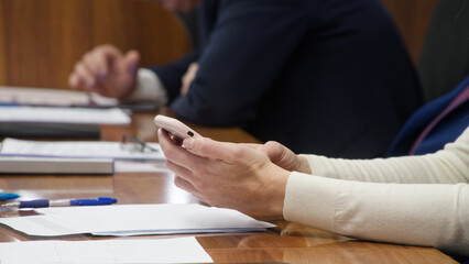 Close up of female hands holding smartphone at wooden conference table during business meeting. Woman texting message or browsing internet while colleague sits in background. Photo