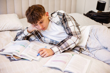 A teenage boy in a plaid shirt lies on his bed and studies with textbooks and notebooks, focusing on his homework. A natural lifestyle scene showing everyday student life and learning at home.