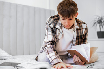 A teenage boy in a plaid shirt lies on his bed and studies with textbooks and notebooks, focusing...