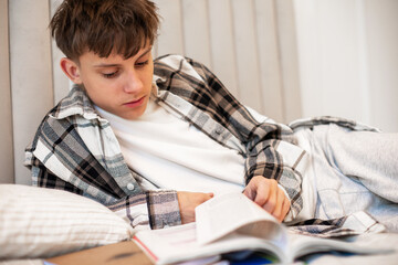 A teenage boy in a plaid shirt lies on his bed and studies with textbooks and notebooks, focusing...