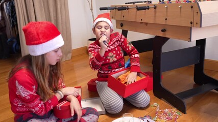 Cute brother and sister wearing santa hats opening their presents on christmas morning. The boy unwraps a karaoke microphone and sings - Powered by Adobe