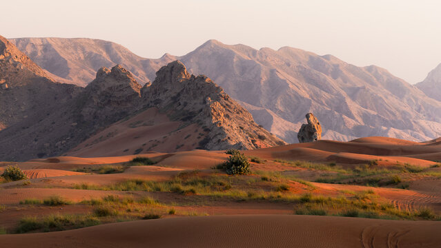 View of sun-kissed sand dunes ripple across the vast desert, meeting rugged mountains under a soft sky, creating a tranquil yet powerful scene, Rub' al-Khali, United Arab Emirates.