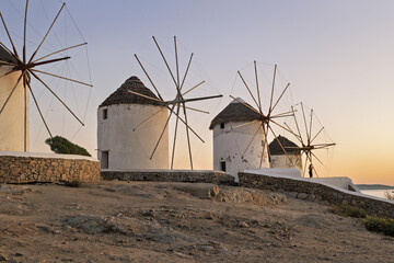 Mykonos, Greece, traditional cylindrical windmills with conical thatched roofs and large wooden blades. The whitewashed structures stand on a stone platform under sunset light.