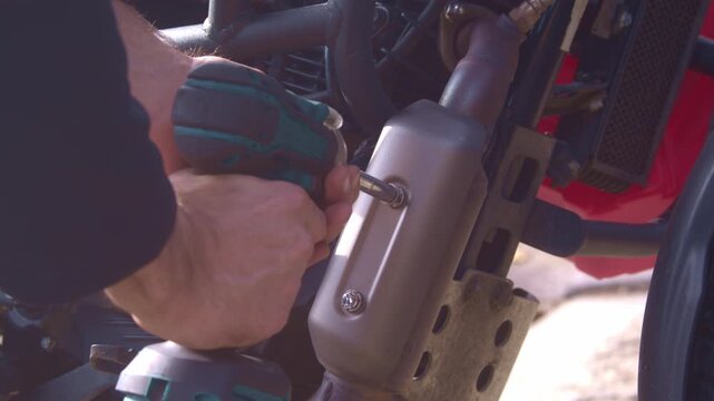 A mechanic repairs a motorcycle outdoors. He's unscrewing a bolt from the muffler resonator cover. He's using a cordless, black-and-blue impact wrench. Over-the-shoulder view from the right.