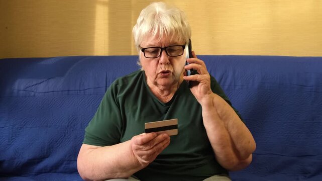 Caucasian elderly woman sitting on sofa holding credit card and talking on phone. Concept of scam, money loss, banking fraud protection, customer service, remote payment