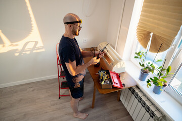 Focused man wearing a headlamp and glasses skillfully uses a soldering iron, repairing electronics at home. Sunlight streams through the window, highlighting the DIY project with tools on a table.