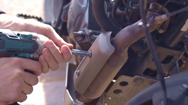A mechanic repairs a motorcycle outdoors. He's unscrewing a bolt from the muffler resonator cover. He's using a cordless, black-and-blue impact wrench, right view.