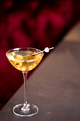 A close-up shot of a classic cocktail, possibly a martini, served in a coupe glass with green olives on a skewer, set against a dark, moody background. The focus is sharp on the glass and olives