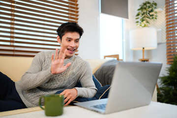 Young adult asian man using a laptop and drinking coffee on a couch in a living room