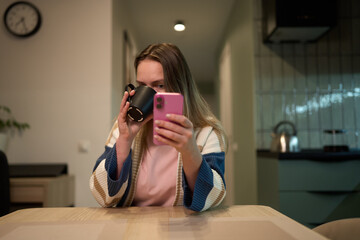 Woman drinking coffee and browsing smartphone at kitchen table