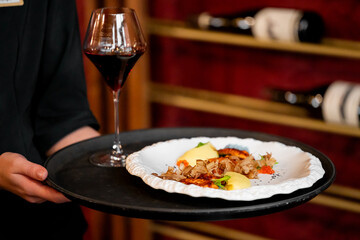 A waiter holds a tray with a gourmet dish and a glass of red wine, with wine bottles blurred in the background of a restaurant or cellar