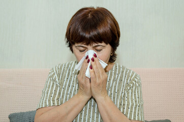 Ill elderly woman sitting on couch and blowing nose into paper tissue and sneezing with closed eyes, cold or flu symptoms, seasonal illness
