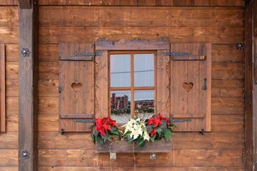 Rustic wooden cabin window with shutters and a flower box of red and white poinsettias. Warm Christmas market detail with heart cut-outs.