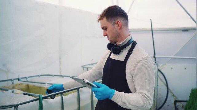 Dutch worker in large agricultural greenhouse measures pH of the water and pH levels in irrigation tank with electronic device. Data is collected on tablet and quality of the irrigation is analyzed.