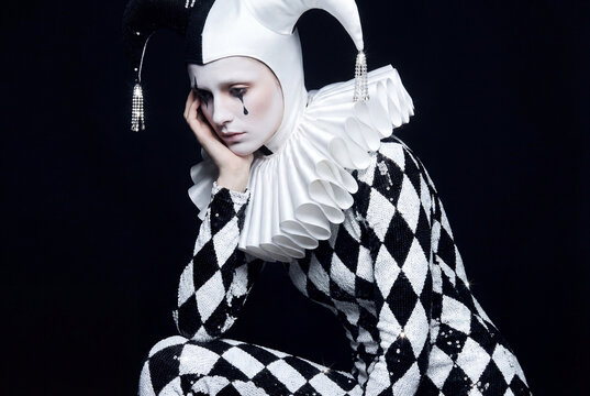 A sad Pierrot or jester in a black and white harlequin costume with a ruff collar, crying a single black tear. Dramatic portrait on a dark background.