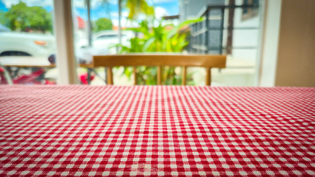 A close-up view of a red and white checkered tablecloth inside a bright café or restaurant. The background is softly blurred, showing a wooden chair, large windows, green plants, and an outdoor area - Powered by Adobe