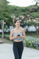 Fototapeta premium Serene Stroll: A radiant woman in athletic wear smiles joyfully as she strolls outdoors, clutching a sleek smartphone in hand, while basking in the embrace of nature.