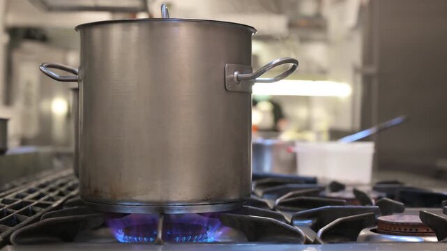 Large stainless-steel pot heating over a blue gas flame in a commercial restaurant kitchen, captured in a static shot during active cooking prep. High quality 4k footage