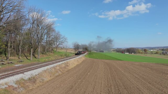 A classic retro steam train chugs along the tracks amid beautiful autumn scenery. The smoke billows from the engine as it moves past fields and trees on a clear, sunny day.