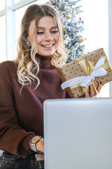 A smiling woman with a gift in her hands chats on a laptop near a festive Christmas tree.
