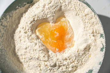 Top-down view of flour with a cracked egg forming a heart-like pool in the center. Concept for baking, love, and cooking at home.