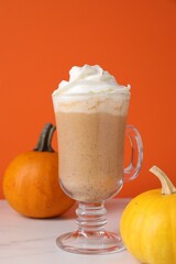 Warm pumpkin latte with whipped cream and fresh vegetables on white marble table against orange background, closeup