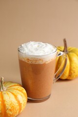 Delicious pumpkin latte with whipped cream in glass cup and fresh vegetables on beige background, closeup