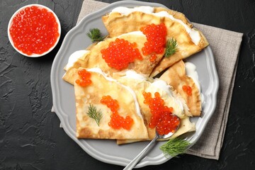 Tasty crepes with red caviar and sour cream on black textured table, flat lay