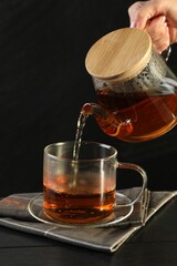 Woman pouring hot tea into cup from teapot at black wooden table, closeup