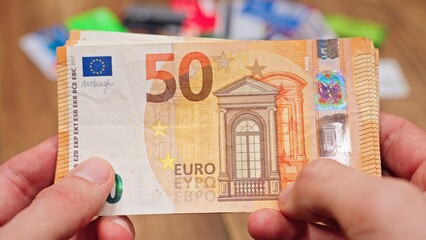 Man holding US hundred dollar bills with both hands, blurred credit cards on a table in the background, representing finances, spending, and cash management.