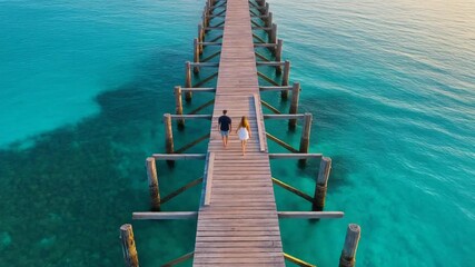 Couple walking on a long wooden pier over turquoise ocean water at sunset, romantic tropical getaway.