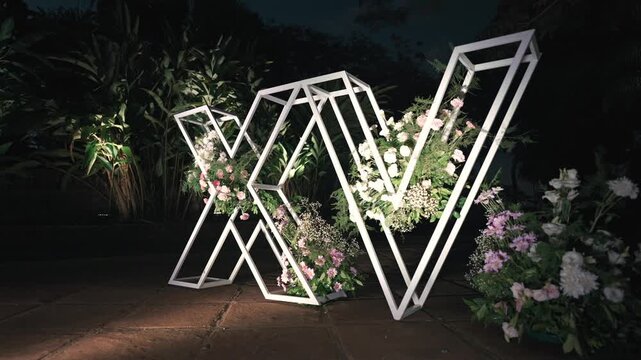 Illuminated metal letters forming &ldquo;XV&rdquo; stand on garden path at night, with open geometric frames filled with pastel flowers, creating modern decorative backdrop for Sweet 15 quincea&ntilde;era celebration