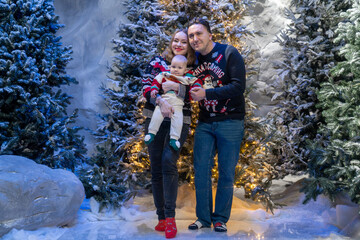family Christmas moment with parents holding baby in snowy holiday setting. Both adults wear festive Christmas sweaters, while baby is dressed in a cute holiday outfit with Santa hat. Snowflakes fall 