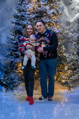 family Christmas moment with parents holding baby in snowy holiday setting. Both adults wear festive Christmas sweaters, while baby is dressed in a cute holiday outfit with Santa hat. Snowflakes fall 