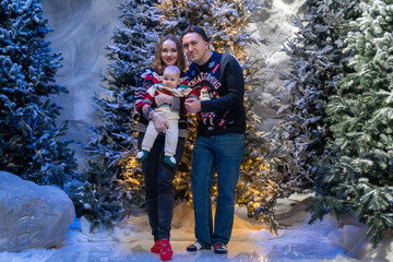 family Christmas moment with parents holding baby in snowy holiday setting. Both adults wear festive Christmas sweaters, while baby is dressed in a cute holiday outfit with Santa hat. Snowflakes fall 