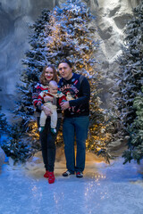 family Christmas moment with parents holding baby in snowy holiday setting. Both adults wear festive Christmas sweaters, while baby is dressed in a cute holiday outfit with Santa hat. Snowflakes fall 