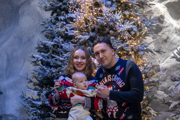 family Christmas moment with parents holding baby in snowy holiday setting. Both adults wear festive Christmas sweaters, while baby is dressed in a cute holiday outfit with Santa hat. Snowflakes fall 