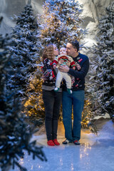 family Christmas moment with parents holding baby in snowy holiday setting. Both adults wear festive Christmas sweaters, while baby is dressed in a cute holiday outfit with Santa hat. Snowflakes fall 