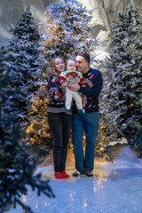 family Christmas moment with parents holding baby in snowy holiday setting. Both adults wear festive Christmas sweaters, while baby is dressed in a cute holiday outfit with Santa hat. Snowflakes fall 