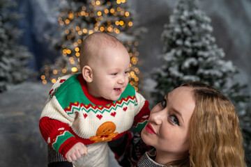 mother lifts her smiling baby in a festive winter scene. The baby wears a colorful Christmas sweater with a gingerbread design, surrounded by snowy trees and warm holiday lights
