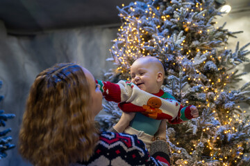 mother lifts her smiling baby in a festive winter scene. The baby wears a colorful Christmas sweater with a gingerbread design, surrounded by snowy trees and warm holiday lights