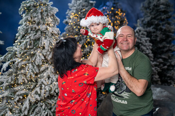 Grandparents posing with grandchild during festive Christmas. baby is dressed in Santa hat and holiday sweater, held lovingly by smiling grandparents. Snow-covered Christmas trees and warm twinkling 