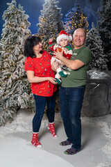 Grandparents posing with grandchild during festive Christmas. baby is dressed in Santa hat and holiday sweater, held lovingly by smiling grandparents. Snow-covered Christmas trees and warm twinkling 