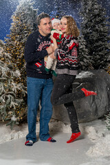Happy family Christmas portrait in snowy winter setting. Mother and father wearing festive holiday sweaters stand together holding baby dressed in a Santa hat and Christmas outfit