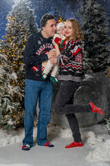 Happy family Christmas portrait in snowy winter setting. Mother and father wearing festive holiday sweaters stand together holding baby dressed in a Santa hat and Christmas outfit