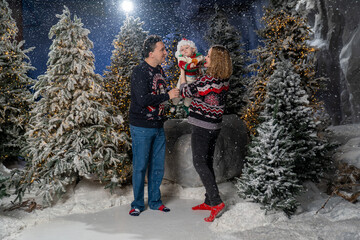 Happy family Christmas portrait in snowy winter setting. Mother and father wearing festive holiday sweaters stand together holding baby dressed in a Santa hat and Christmas outfit