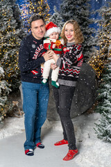 Happy family Christmas portrait in snowy winter setting. Mother and father wearing festive holiday sweaters stand together holding baby dressed in a Santa hat and Christmas outfit