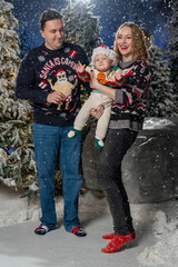 Happy family Christmas portrait in snowy winter setting. Mother and father wearing festive holiday sweaters stand together holding baby dressed in a Santa hat and Christmas outfit